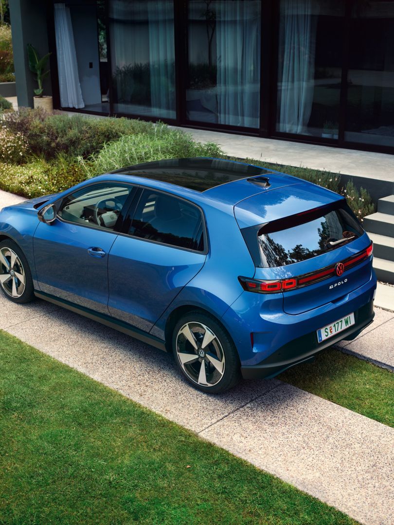 Син VW ID. Polo Style е паркиран пред зелена жилищна сграда, мъж върви зад автомобила.