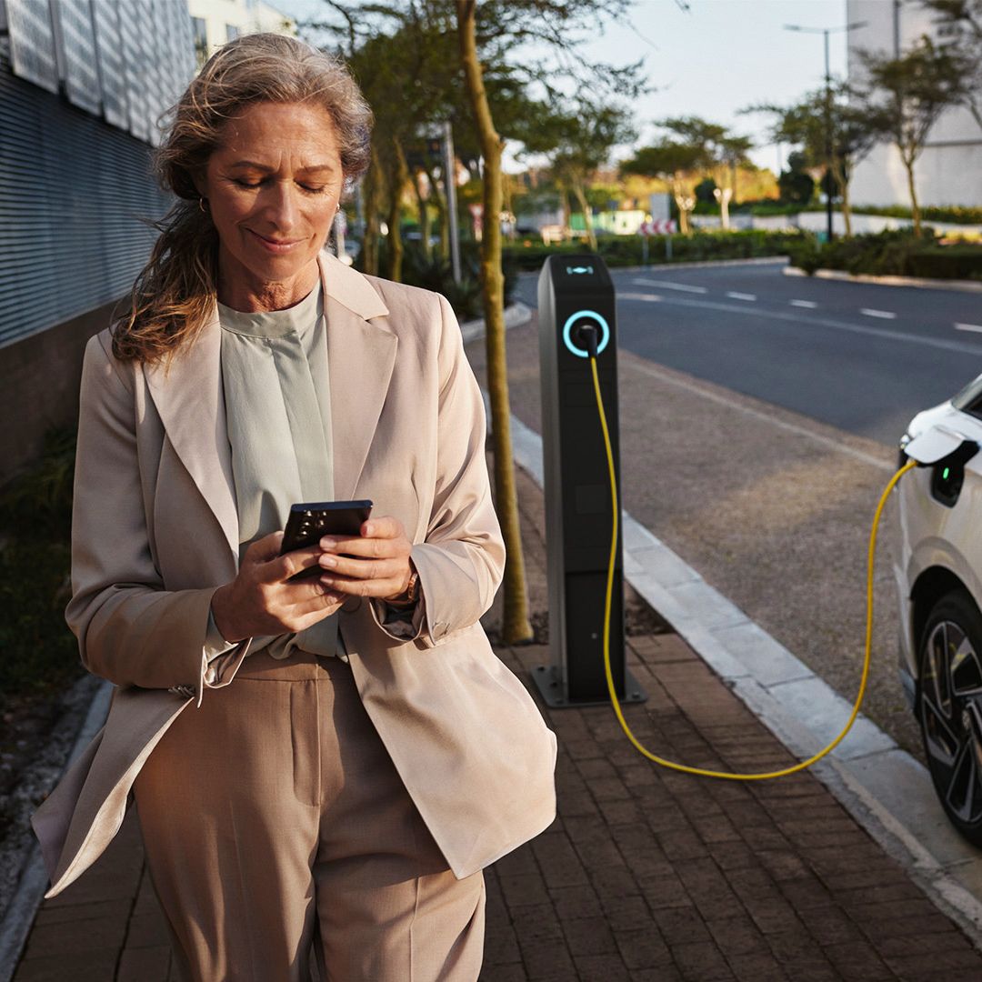 Weißer VW ID. seitlich lädt an einer öffentlichen Ladesäule. Eine Frau geht mit dem Smartphone in der Hand daran vorbei.