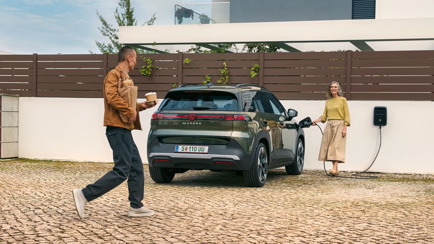 Жена включва кабел за зареждане от стенна кутия в зелен VW ID. Cross, мъж, се приближава до нея с чаши за кафе в ръка и хляб под мишница.