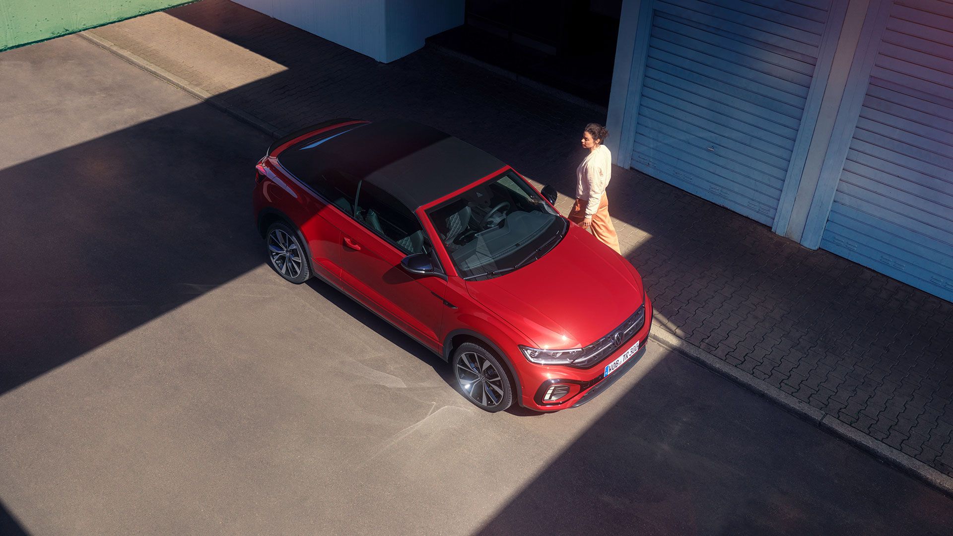Птичи поглед към VW T-Roc Cabriolet в червен цвят с отворен покрив. Паркиран отстрани на пътя, жена минава покрай автомобила. 
