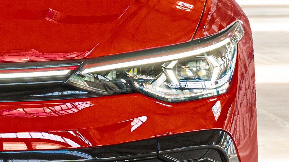 Nahaufnahme eines roten Golf GTI, die sich auf einen Frontscheinwerfer konzentriert. Die Oberfläche ist glänzend und reflektiert das Licht. Der Scheinwerfer hat ein modernes, scharfkantiges Design.