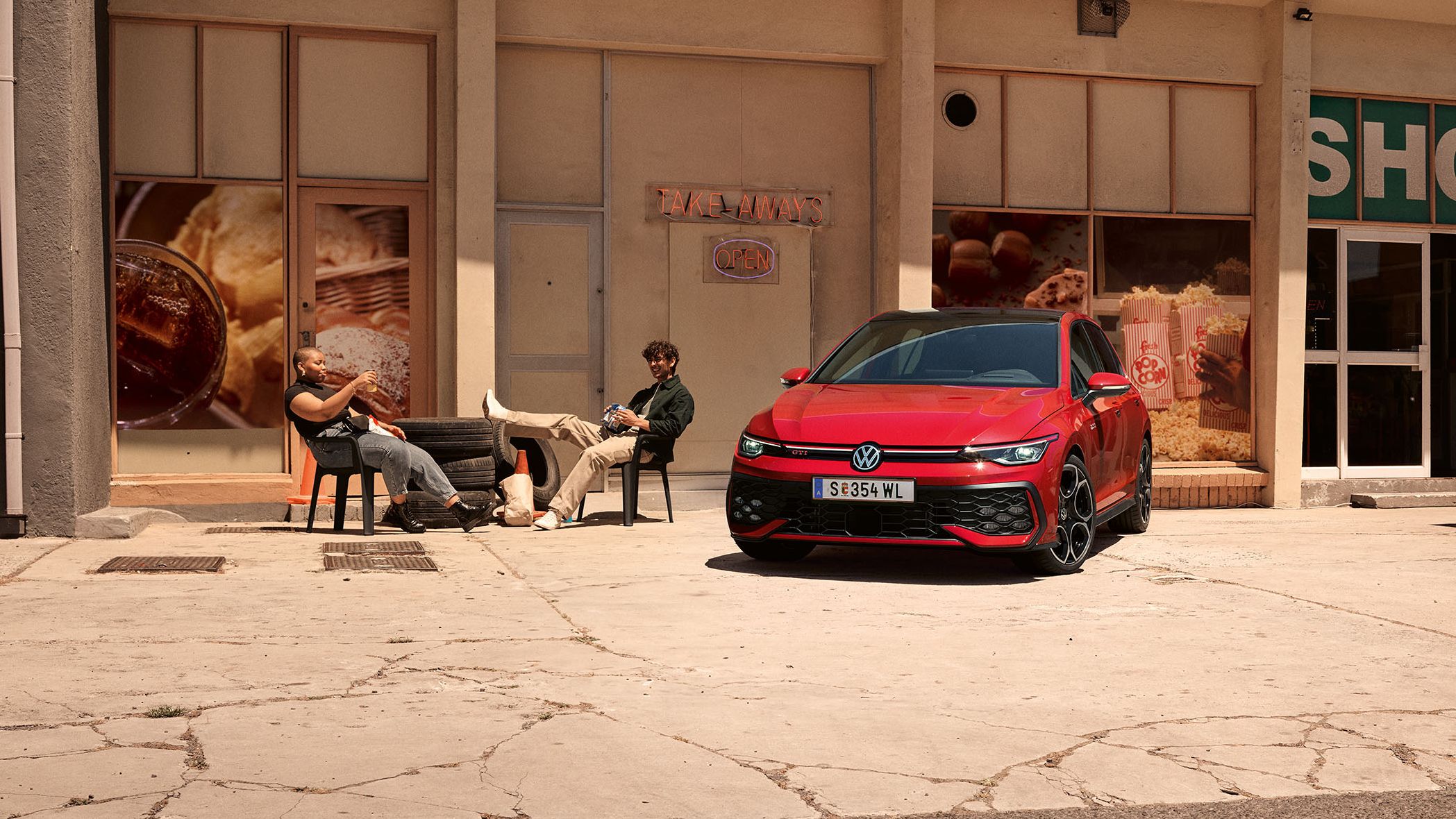 Zwei Personen sitzen auf Plastikstühlen neben einem Golf GTI, dahinter befindet sich ein Shop.