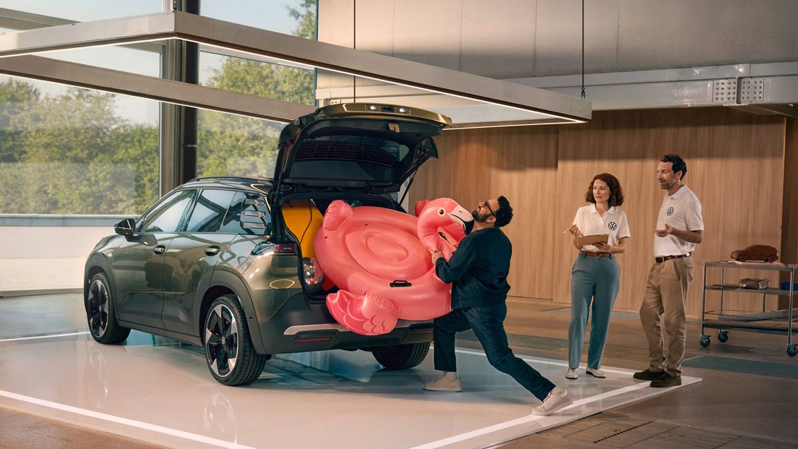 Мъж бута надуваемо фламинго в отворения багажник на VW ID. Кръст. Двама души стоят до него и го наблюдават с усмивка.