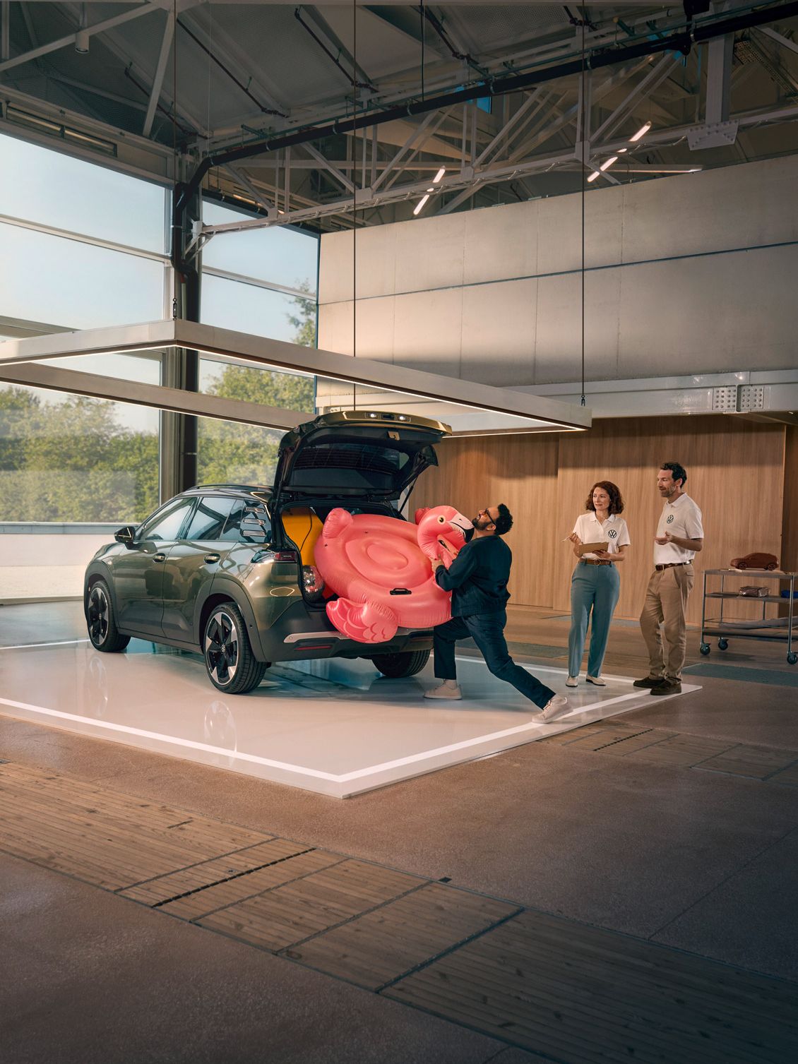 Мъж бута надуваемо фламинго в отворения багажник на VW ID. Кръст. Двама души стоят до него и го наблюдават с усмивка.