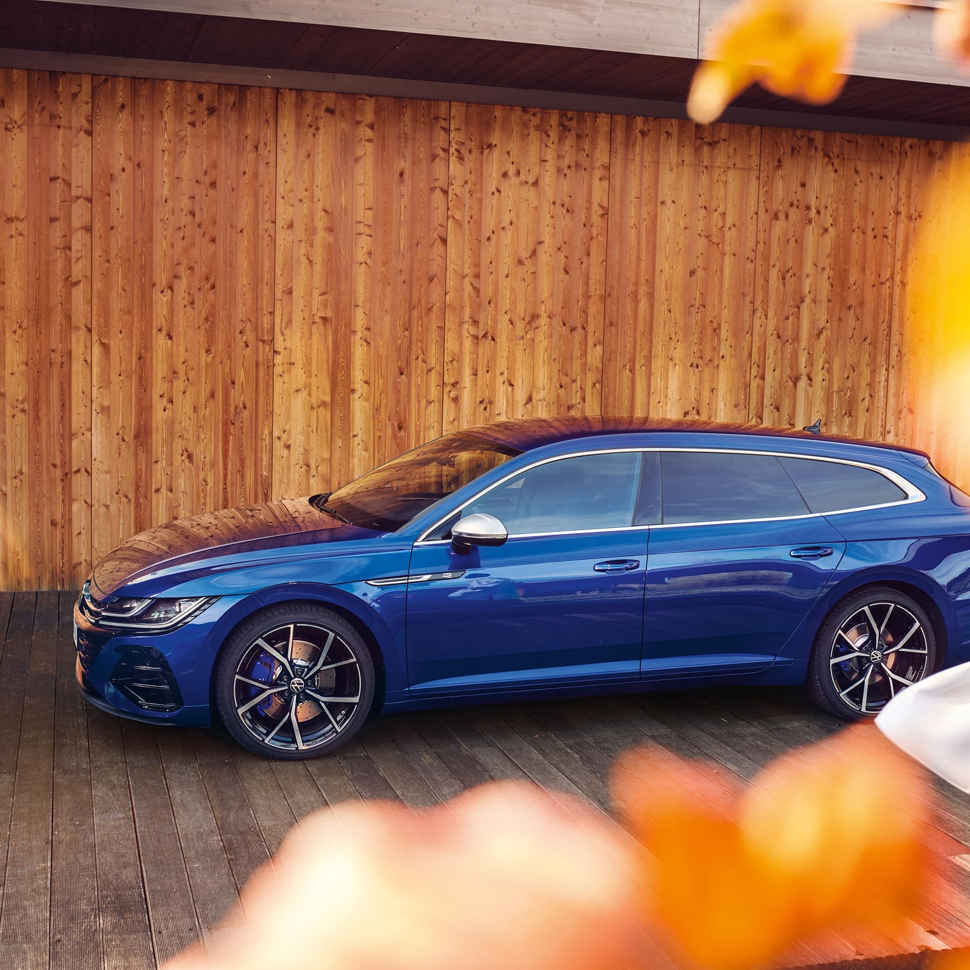 Елегантен син Volkswagen Arteon Shooting Brake паркиран пред модерна дървена сграда с есенни акценти