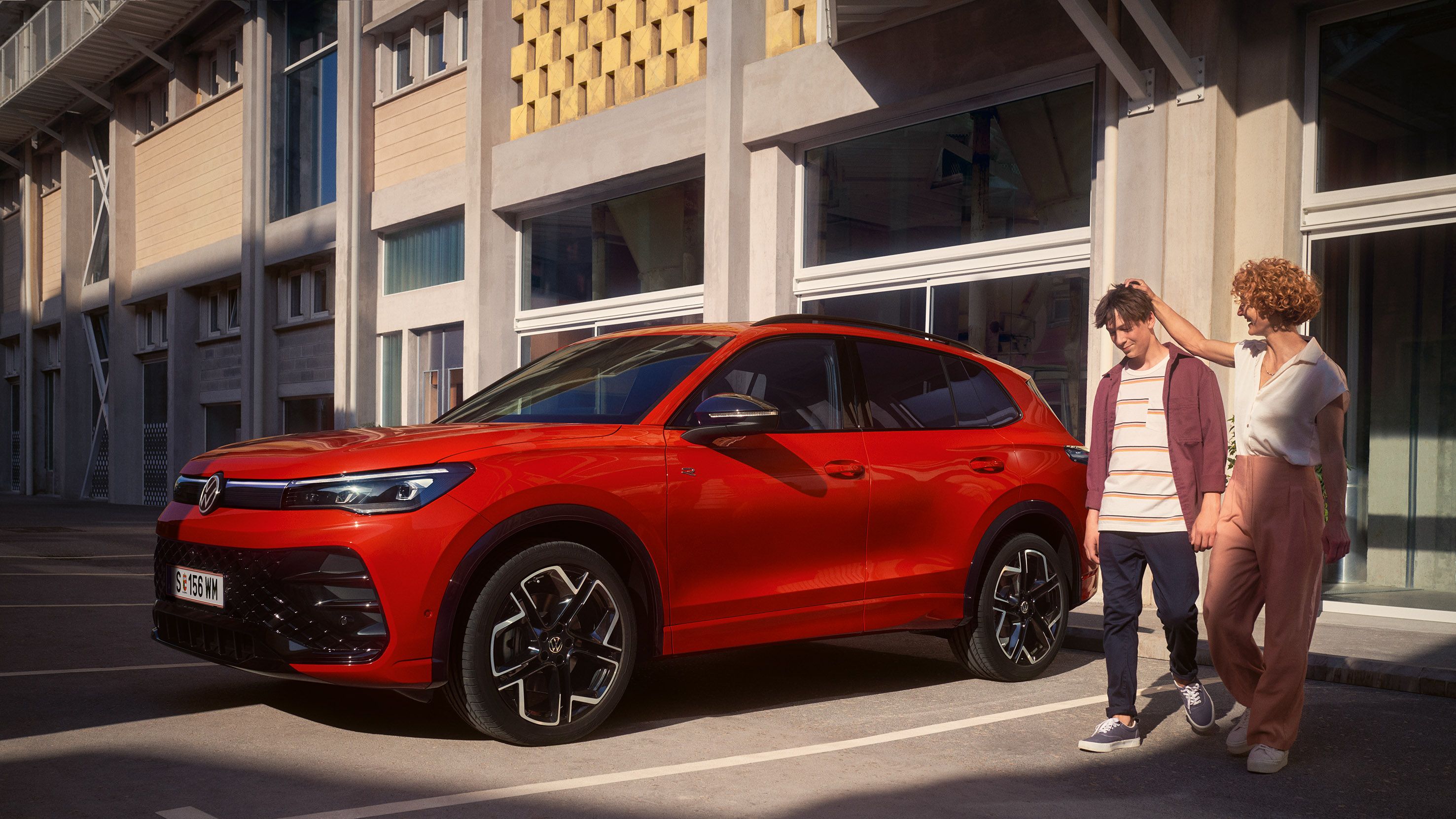 Червен VW Tiguan е паркиран към сграда, към него приближават жена и младеж.