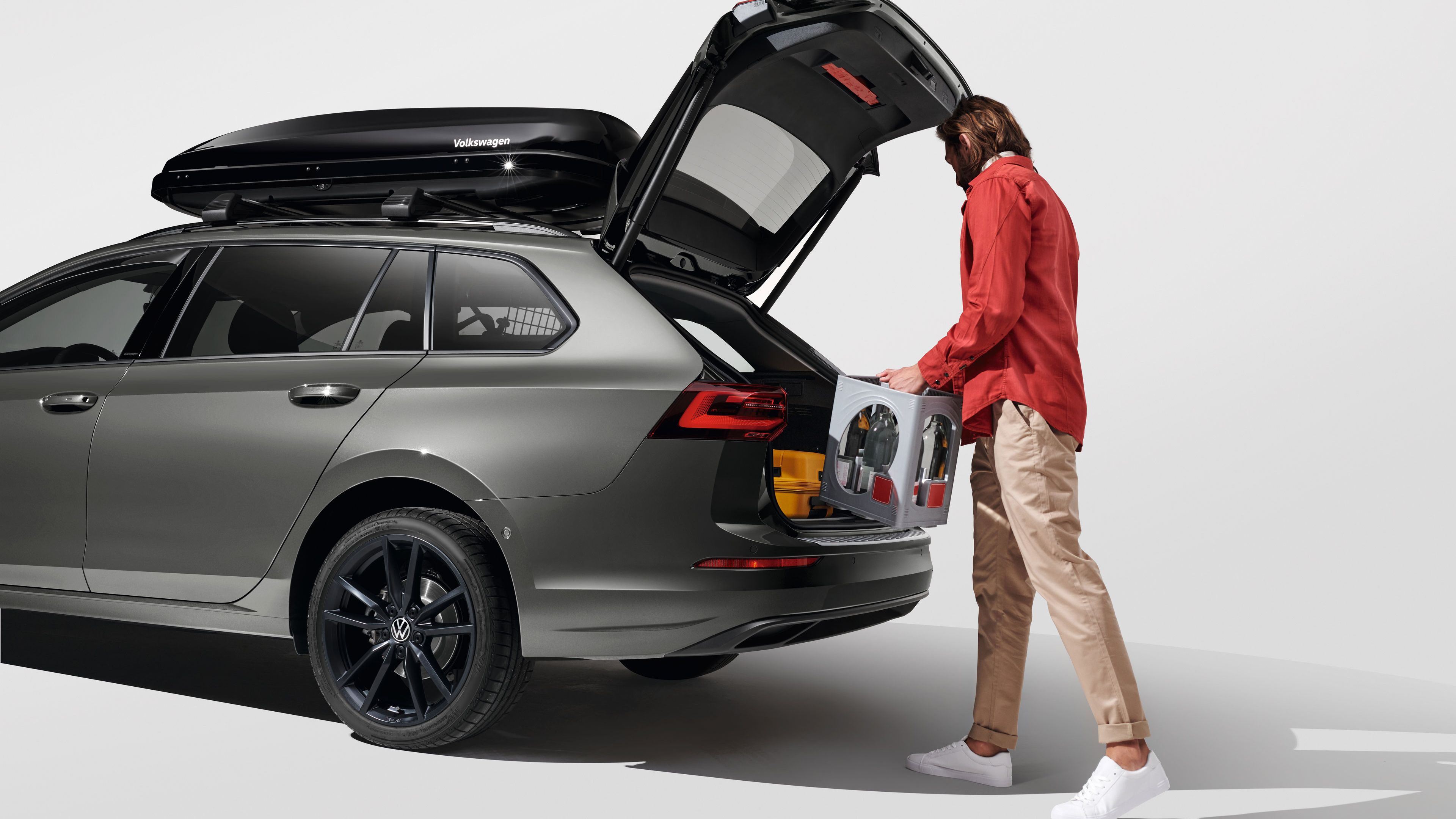 Мъж, облечен в oранжево яке, товарещ багаж в багажника на сив Volkswagen Golf Variant. Автомобилът има товарен багажник на покрива. Той стои на бял фон.