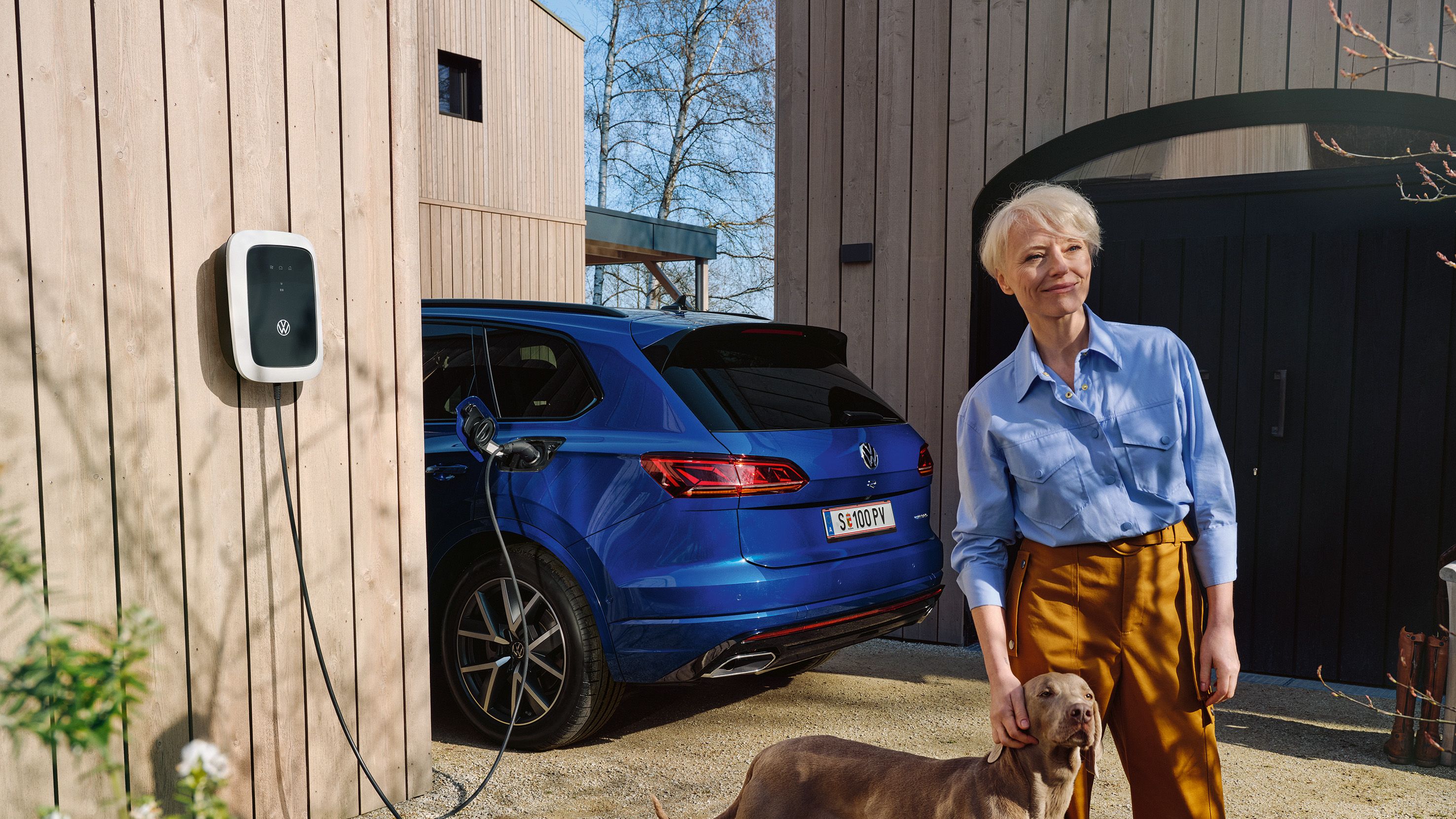 Eine Frau steht mit ihrem Hund vor einem ladenden VW Touareg R eHybrid.
