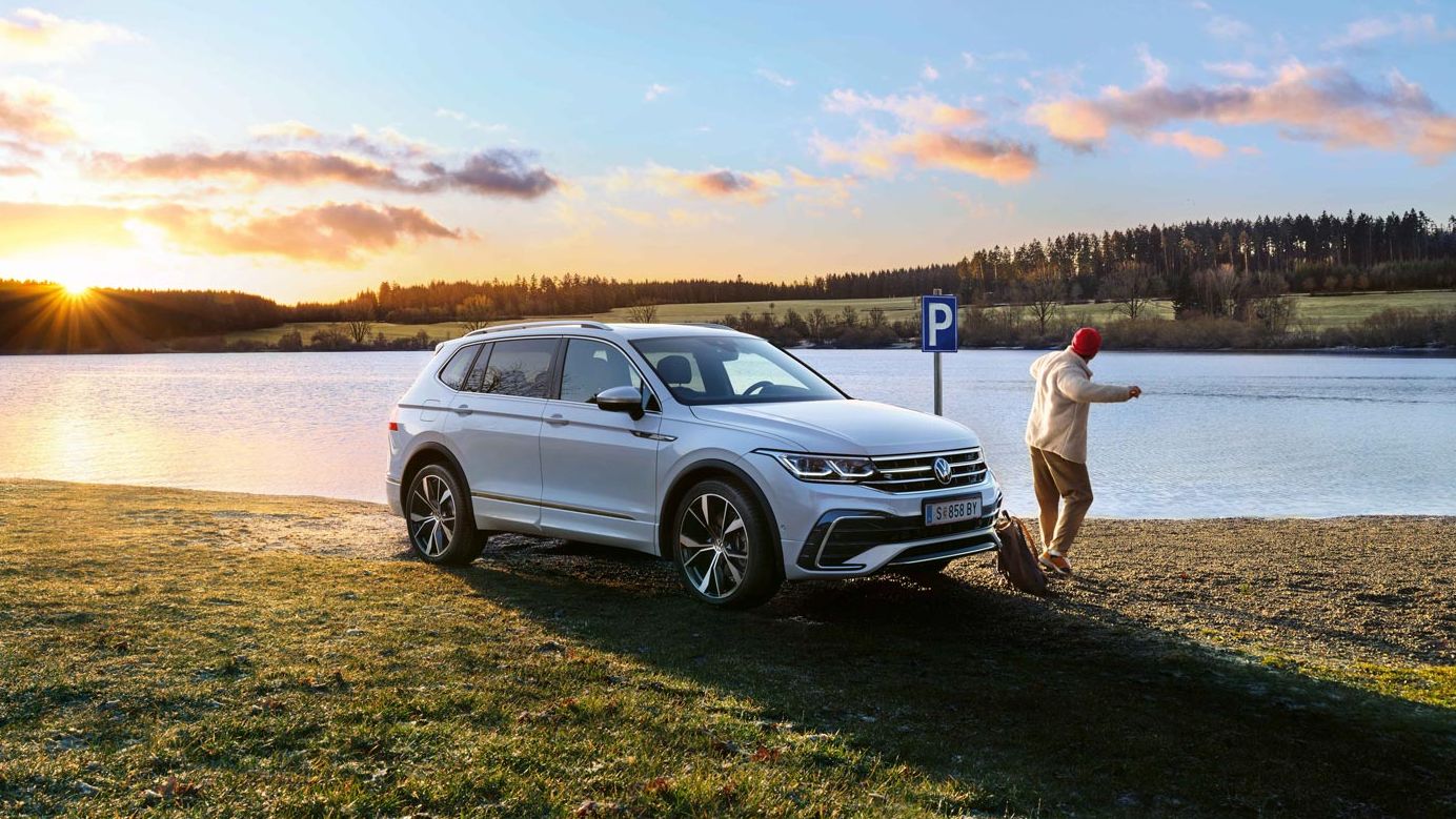 Бял Volkswagen Tiguan паркиран край езеро по залез с човек, който се разхожда наблизо.