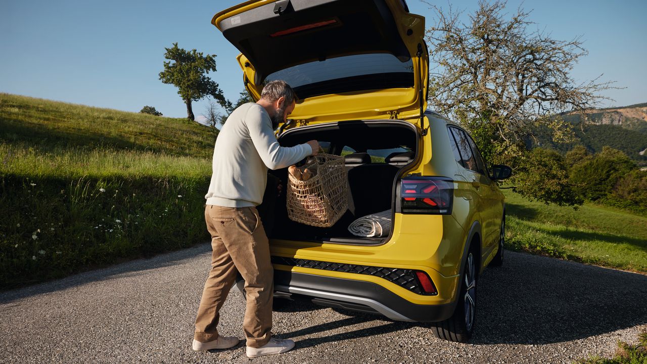 Мъж поставя голяма кошница за пикник в багажника на жълт VW T-Cross на пътя сред хълмист пейзаж.