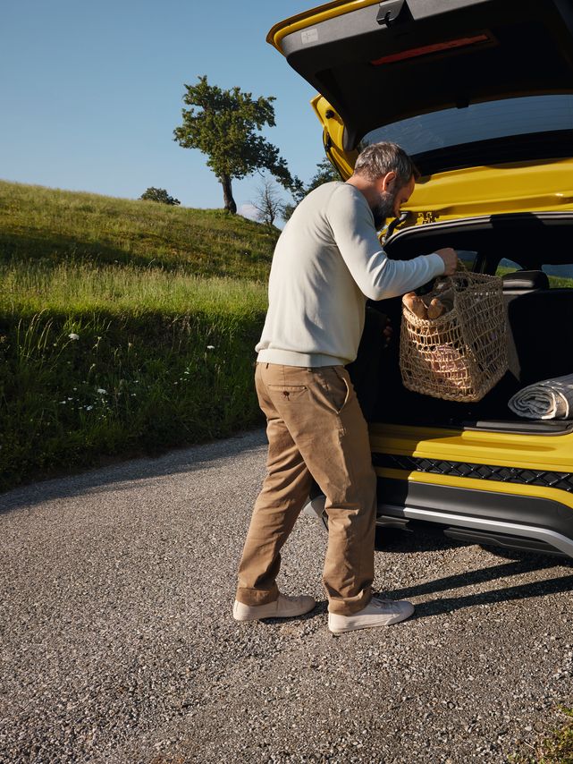 Мъж поставя голяма кошница за пикник в багажника на жълт VW T-Cross на пътя сред хълмист пейзаж.