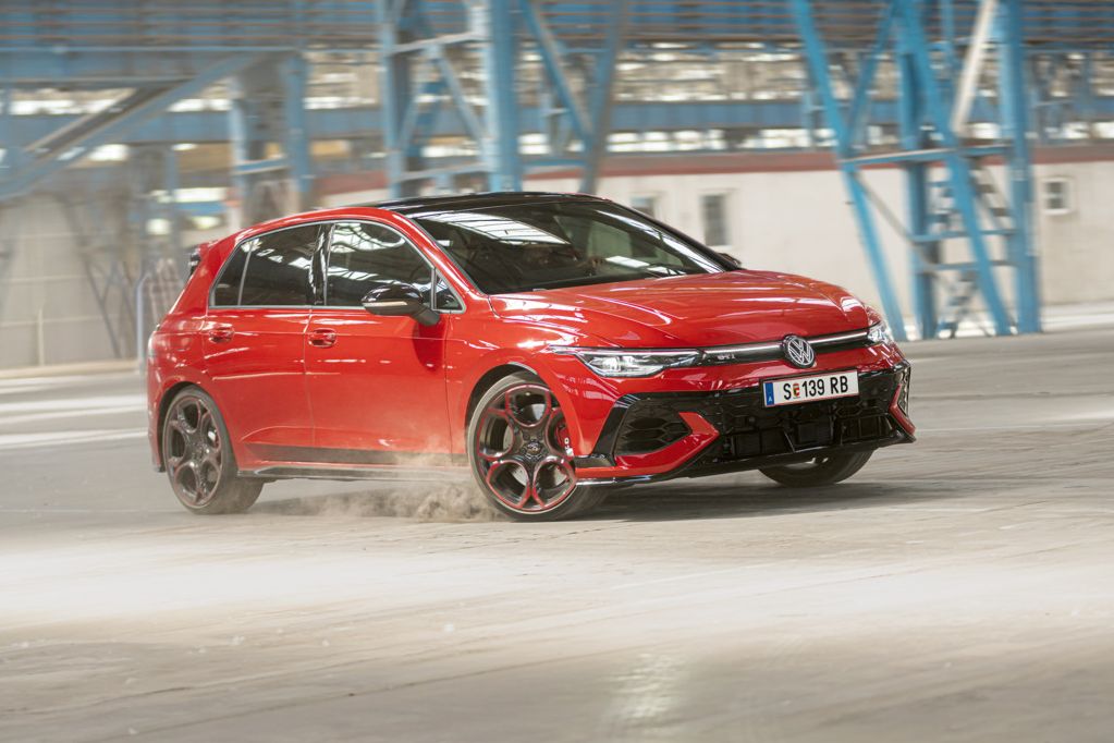 Ein roter Volkswagen Golf GTI fährt schnell durch eine leere Industriehalle. Staub wird von den Reifen aufgewirbelt. Im Hintergrund sind blaue Stahlkonstruktionen und Fenster zu sehen.