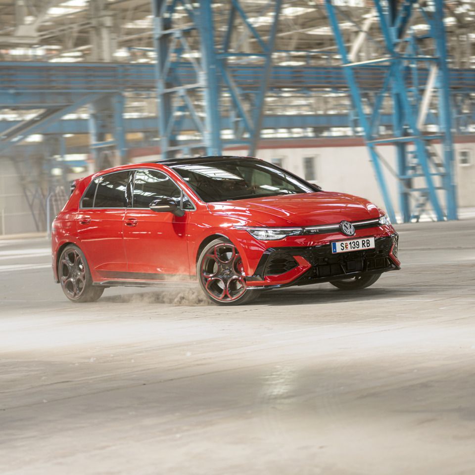 Ein roter Volkswagen Golf GTI fährt schnell durch eine leere Industriehalle. Staub wird von den Reifen aufgewirbelt. Im Hintergrund sind blaue Stahlkonstruktionen und Fenster zu sehen.