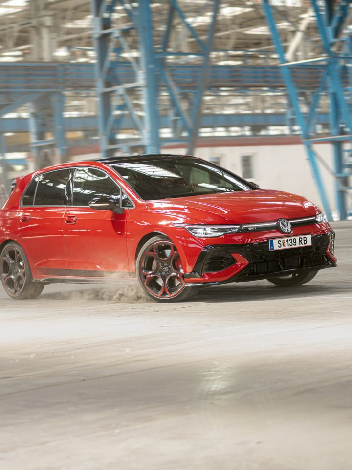 Ein roter Volkswagen Golf GTI fährt schnell durch eine leere Industriehalle. Staub wird von den Reifen aufgewirbelt. Im Hintergrund sind blaue Stahlkonstruktionen und Fenster zu sehen.