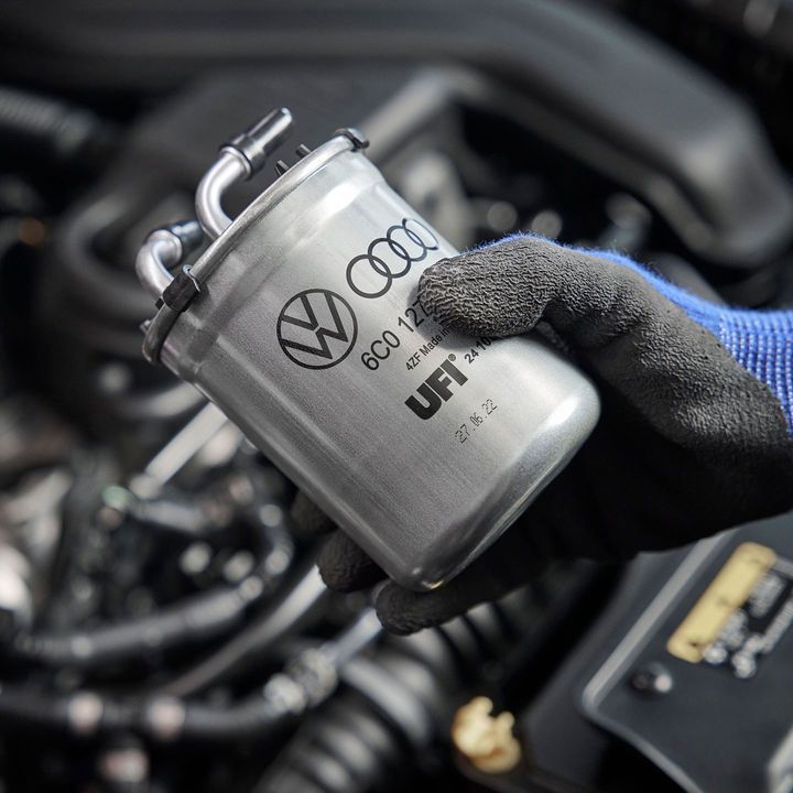 Механик държи сребърен автомобилен филтър, обозначен с логото на Volkswagen, пред автомобилен двигател. Фонът показва части от двигателя и кабели. Механикът носи ръкавици, осигуряващи защита.