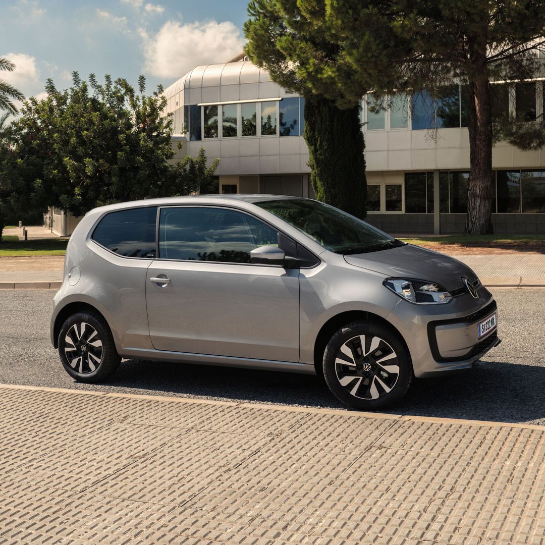 Сребрист Volkswagen e-up! паркиран на улица пред модерна сграда и зеленина