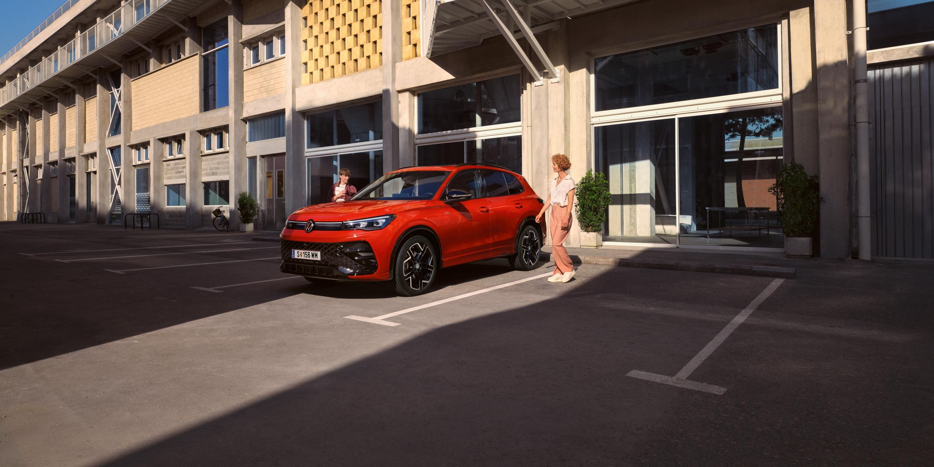 Червен VW Tiguan в профил е паркиран пред сграда, млада жена отива към шофьорското място.
