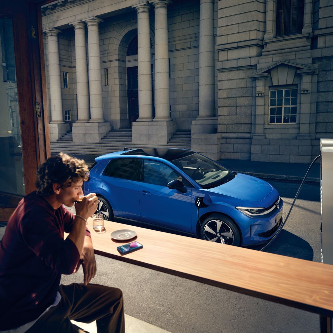 Мъж седи в кафене, през отворен прозорец се вижда синьото Volkswagen ID. Polo Style, което се зарежда на зарядна станция.