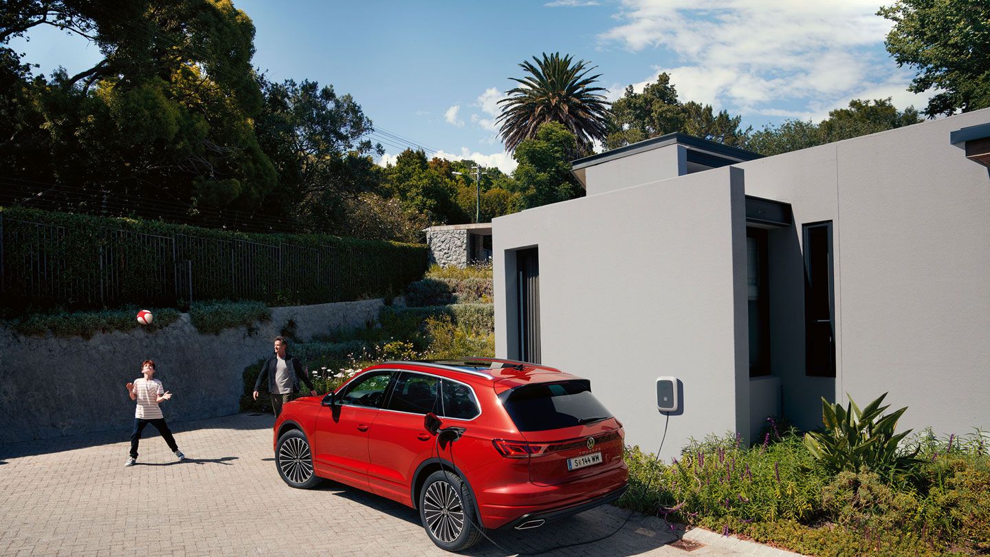 Изглед в ¾ отзад на VW Touareg Elegance eHybrid, включен в зарядна станция пред модерен дом.