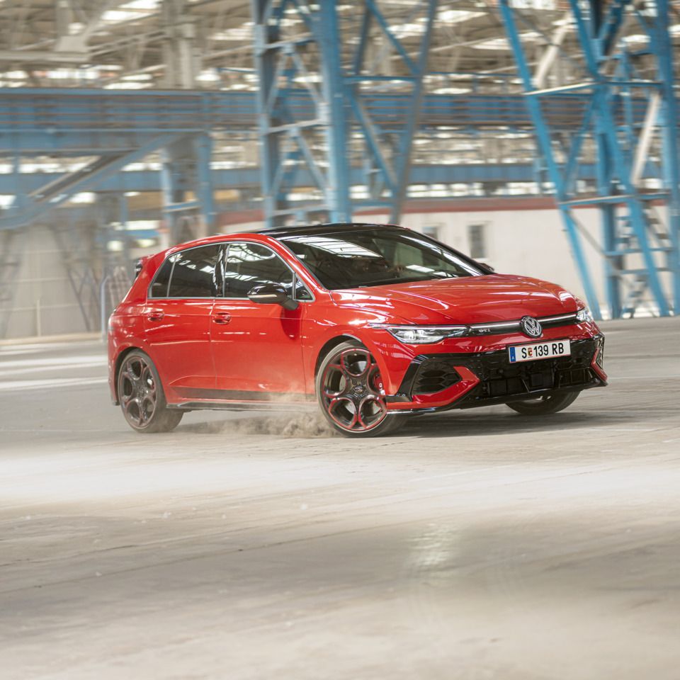 Ein roter Volkswagen Golf GTI fährt schnell durch eine leere Industriehalle. Staub wird von den Reifen aufgewirbelt. Im Hintergrund sind blaue Stahlkonstruktionen und Fenster zu sehen.