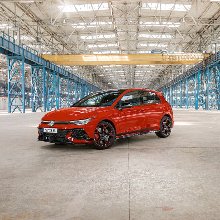 Ein roter Golf GTI steht in einer großen, leeren Industriehalle mit blauen Stahlträgern. Das Dach der Halle ist aus Glas und lässt viel Licht herein.