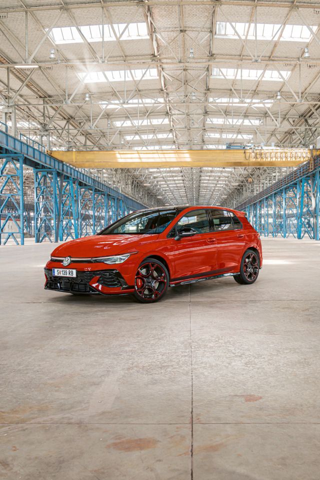 Ein roter Golf GTI steht in einer großen, leeren Industriehalle mit blauen Stahlträgern. Das Dach der Halle ist aus Glas und lässt viel Licht herein.