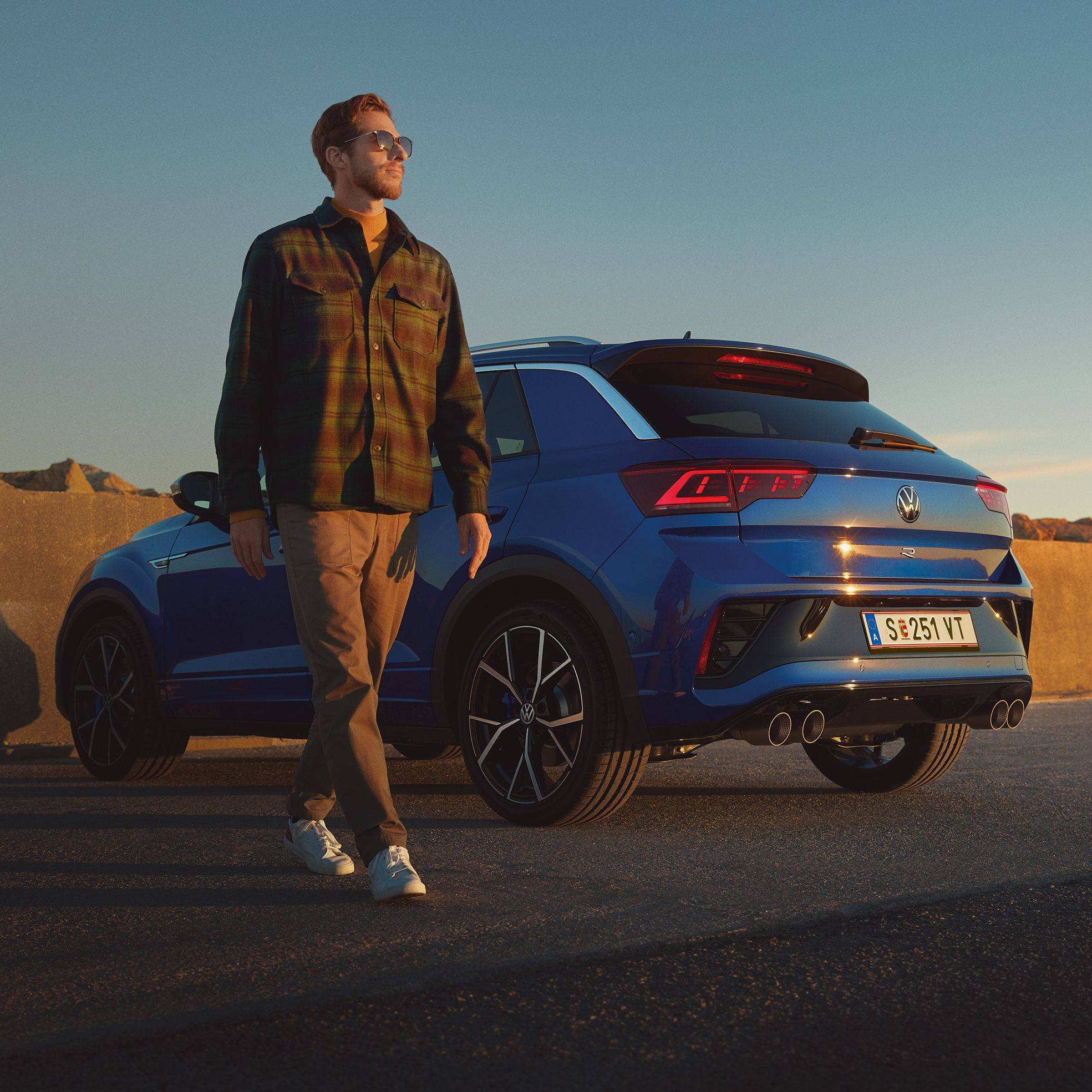 Син Volkswagen T-Roc R, паркиран на крайбрежен път при залез с мъж, който стои до автомобила.