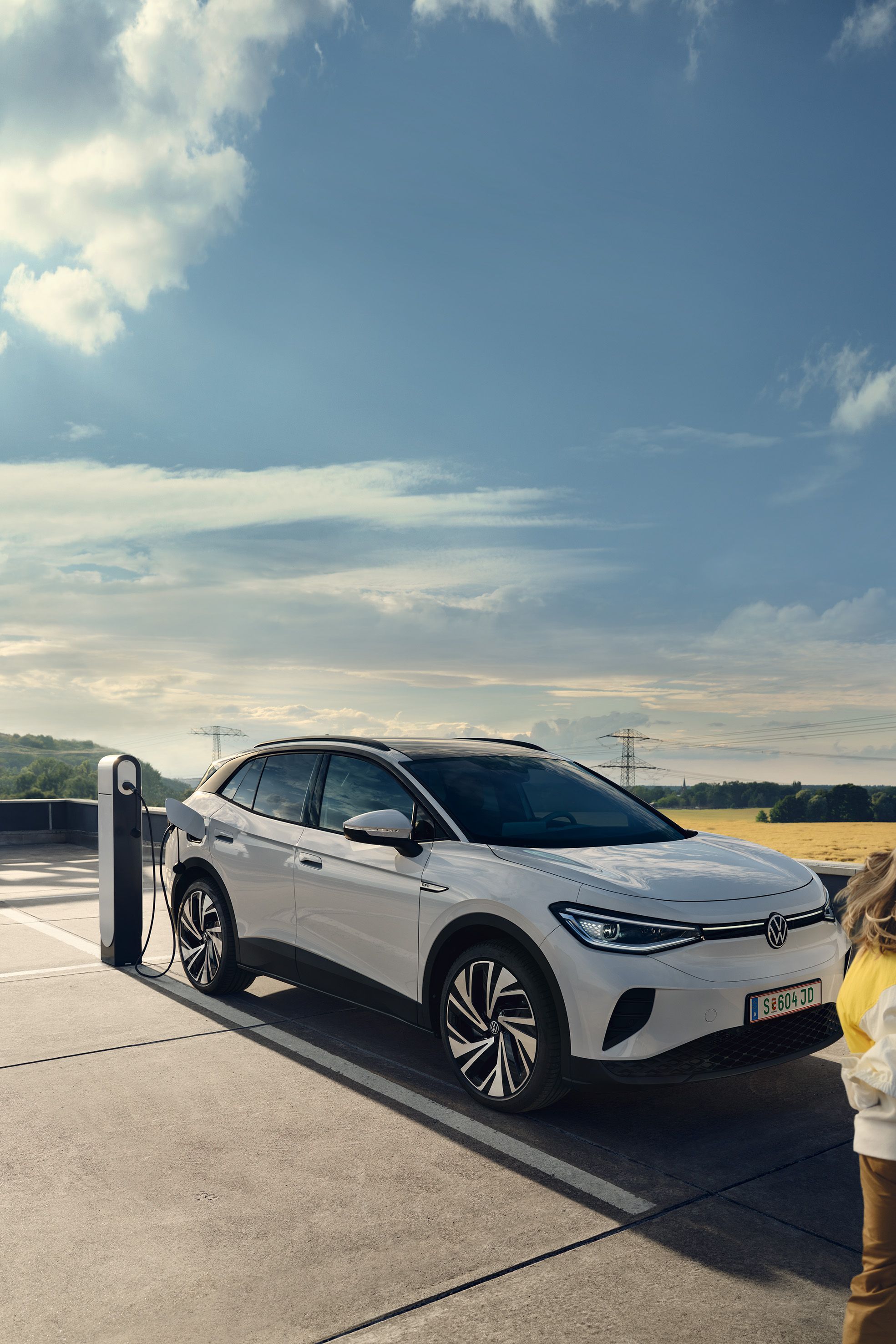 Бял  VW ID.4 се зарежда на зарядна колонка, покрай него преминава дете.