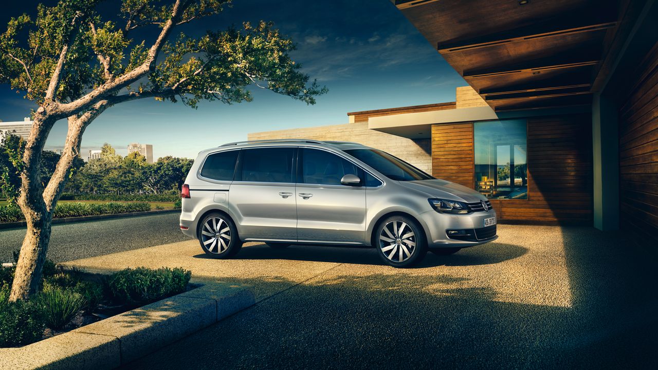 Сребрист Volkswagen Sharan, паркиран в двора на съвременна къща с панорамна гледка към града.