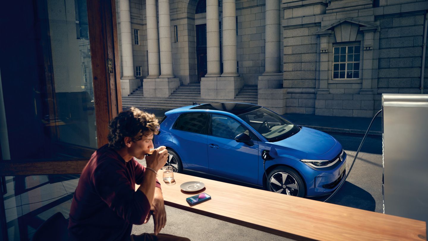 Мъж седи в кафене, през отворен прозорец се вижда синьото Volkswagen ID. Polo Style, което се зарежда на зарядна станция.