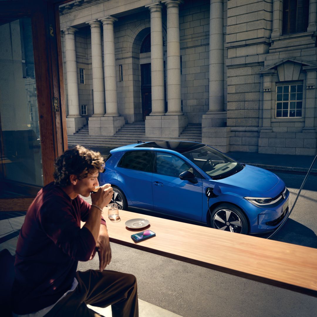 Мъж седи в кафене, през отворен прозорец се вижда синьото Volkswagen ID. Polo Style, което се зарежда на зарядна станция.