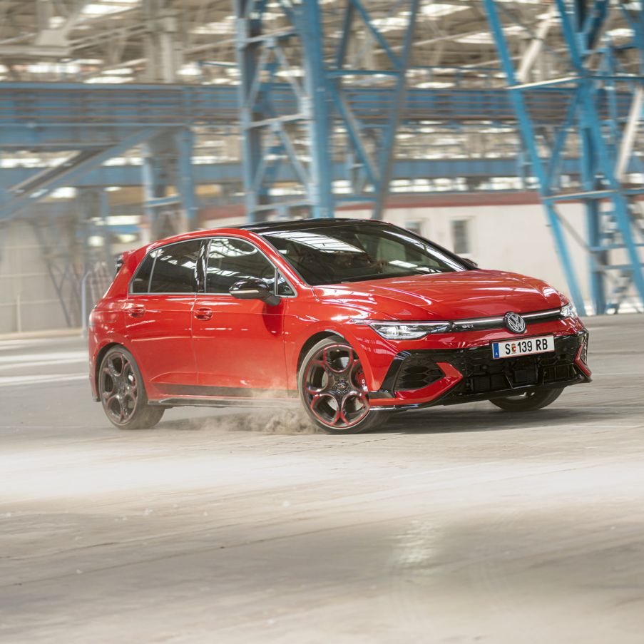 Ein roter Volkswagen Golf GTI fährt schnell durch eine leere Industriehalle. Staub wird von den Reifen aufgewirbelt. Im Hintergrund sind blaue Stahlkonstruktionen und Fenster zu sehen.