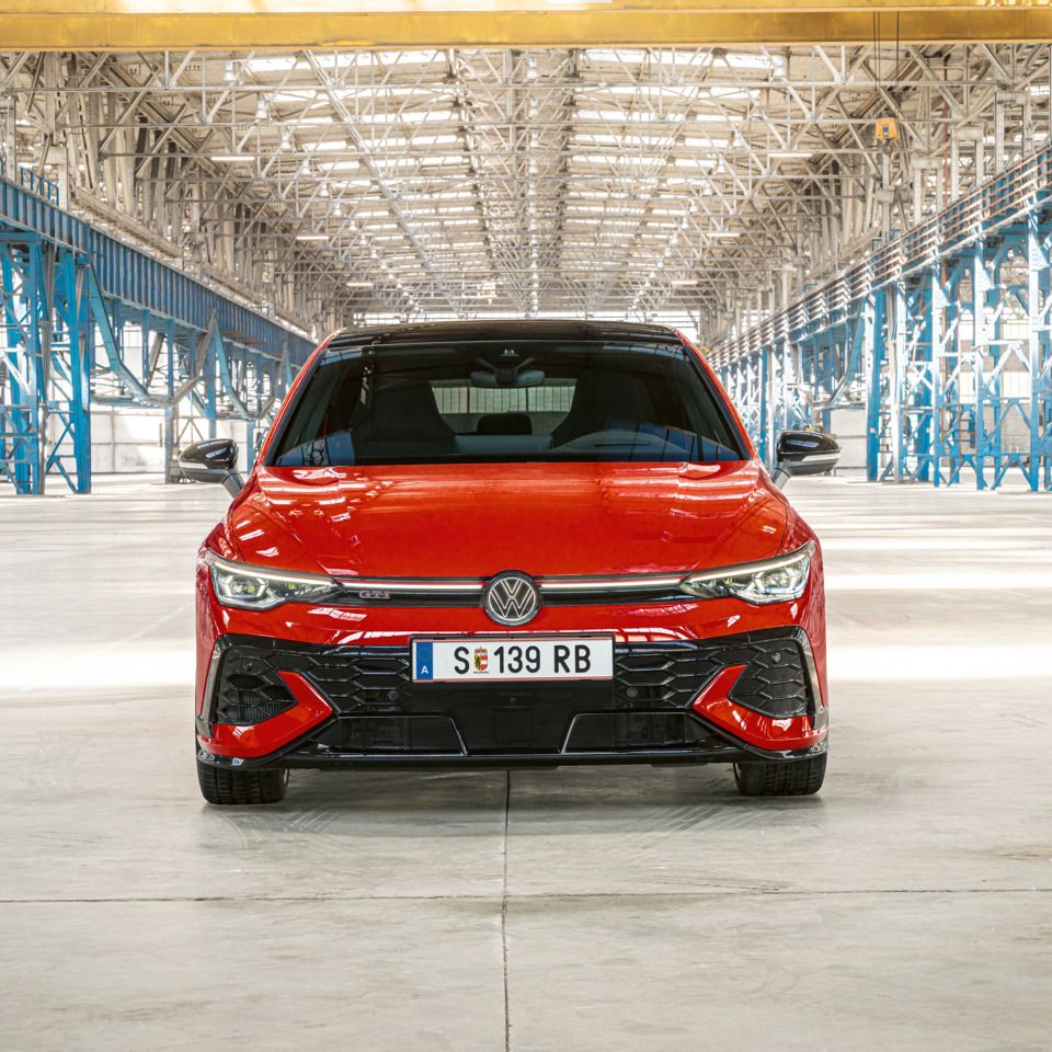 Ein roter Golf GTI steht in einer großen, hell beleuchteten Lagerhalle. Das Auto ist von vorne zu sehen und hat das Kennzeichen MÖB-GT 150. Im Hintergrund sind metallene Strukturen und Säulen zu sehen.