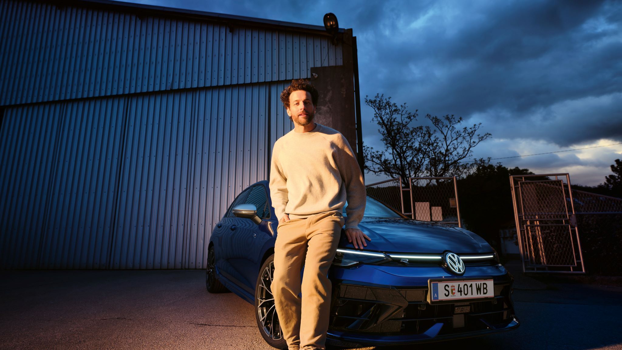 Mann lehnt sich an die Frontseite vom Golf R an, welcher vor einer Halle geparkt ist.