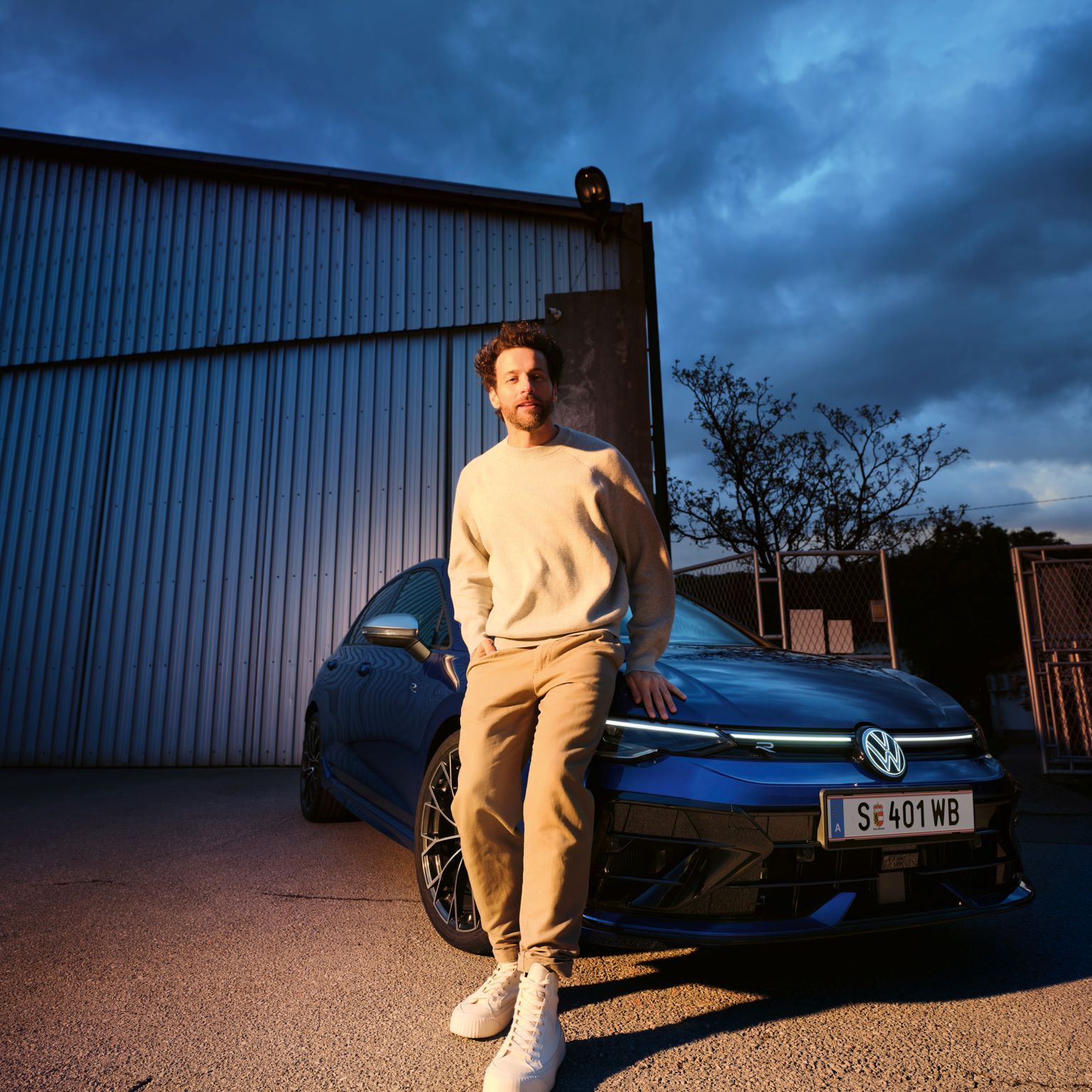 Mann lehnt sich an die Frontseite vom Golf R an, welcher vor einer Halle geparkt ist.