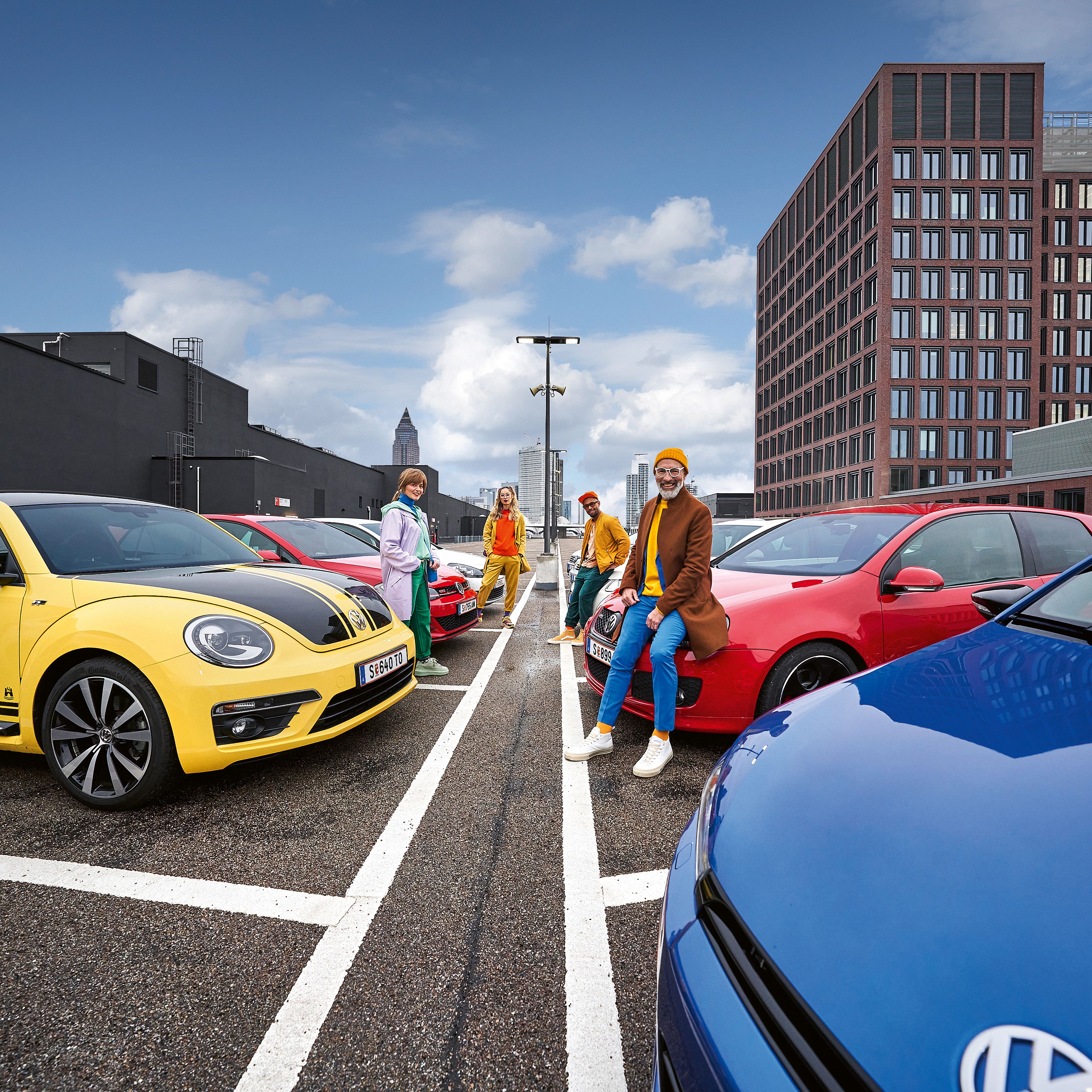 Малка групичка от хора, заобиколени от различни VW модели, са се облегнали на тях. Жълт Beetle, Golf GTI различни моделни генерации и други. В даличината се виждат бизнес сгради.
