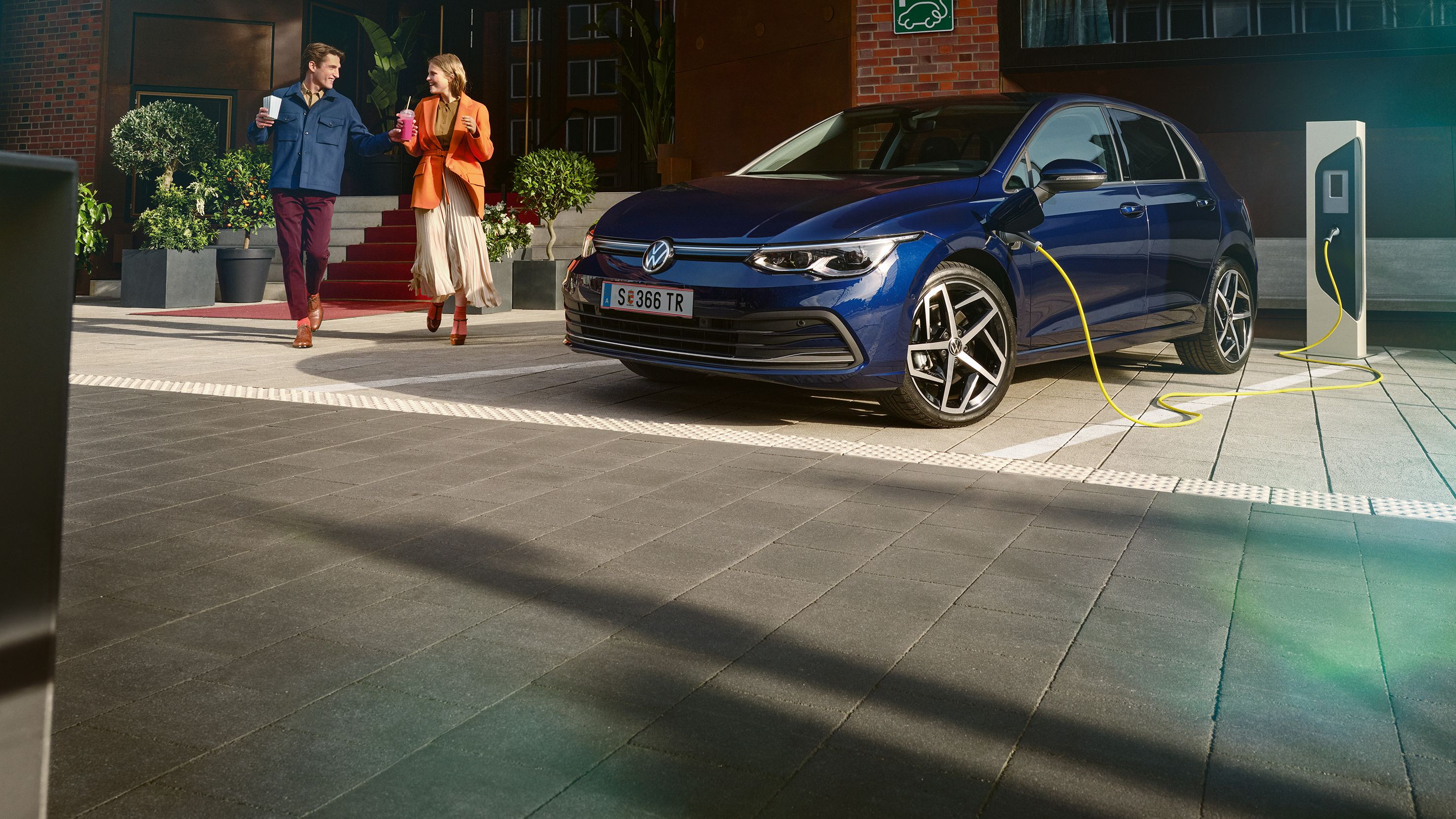 Млада двойка с кафе преминава покрай син Volkswagen Golf GTE, който се зарежда на електрическа станция