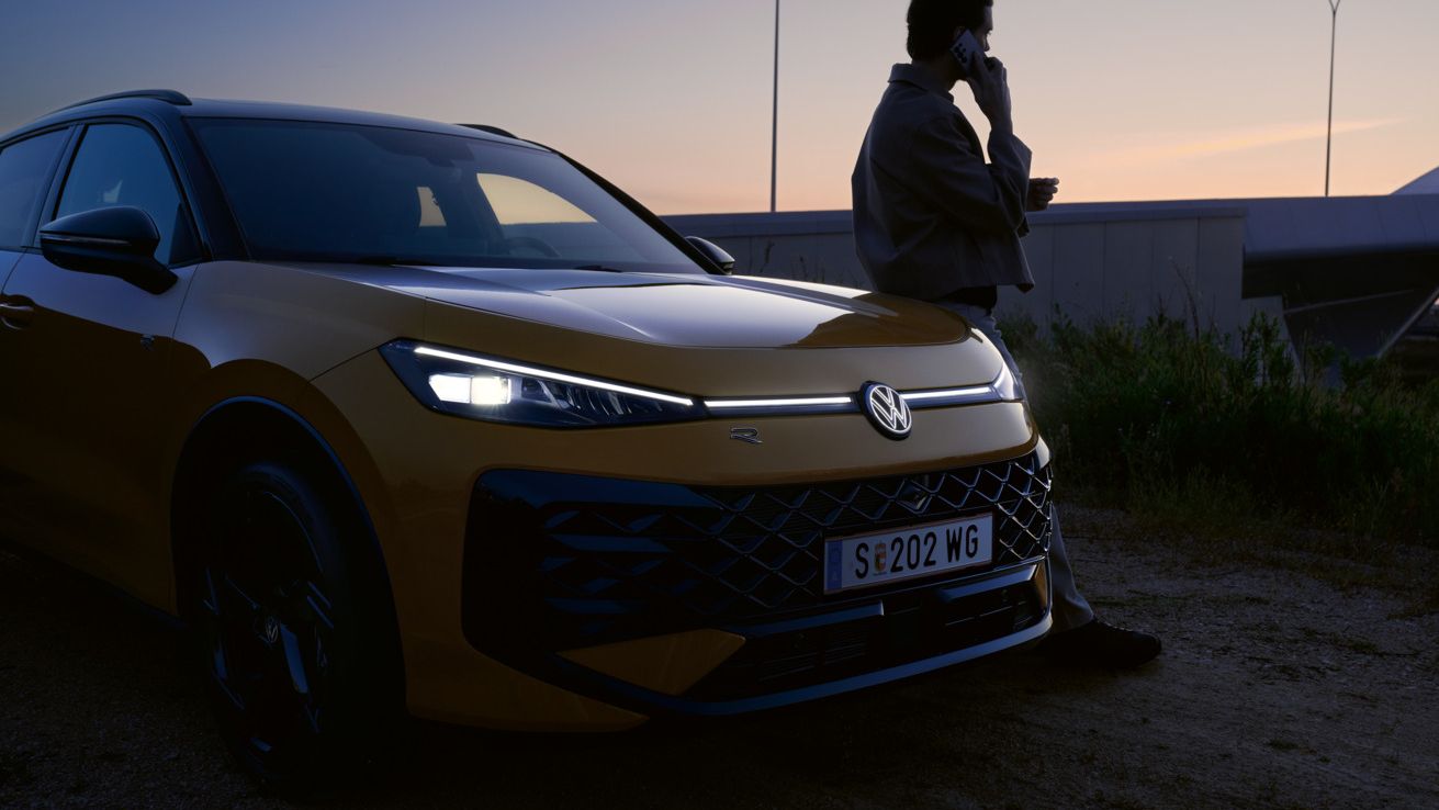 Жълт Volkswagen T-Roc R-Line в здрач с включени предни светлини и силует на мъж до автомобила.