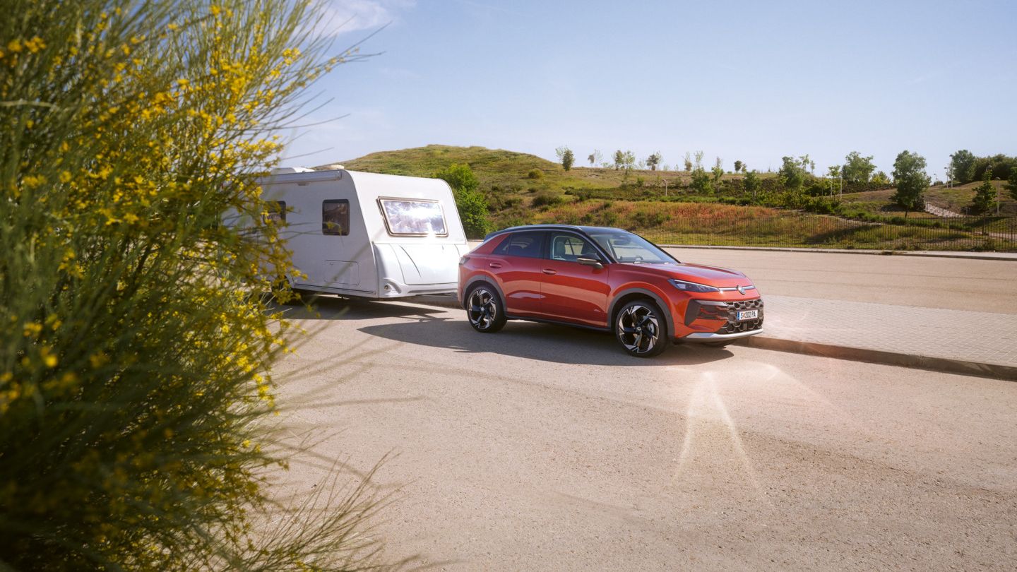 Червен Volkswagen T-Roc тегли каравана на открит път сред зелена природа.