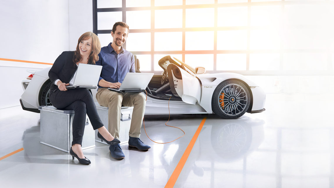 Мъж и жена седят пред Porsche с лаптоп 