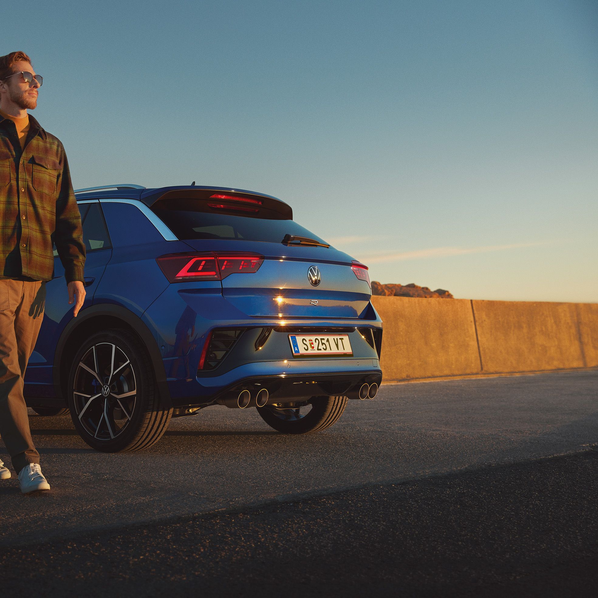 Син Volkswagen T-Roc R, паркиран на крайбрежен път при залез с мъж, който стои до автомобила.