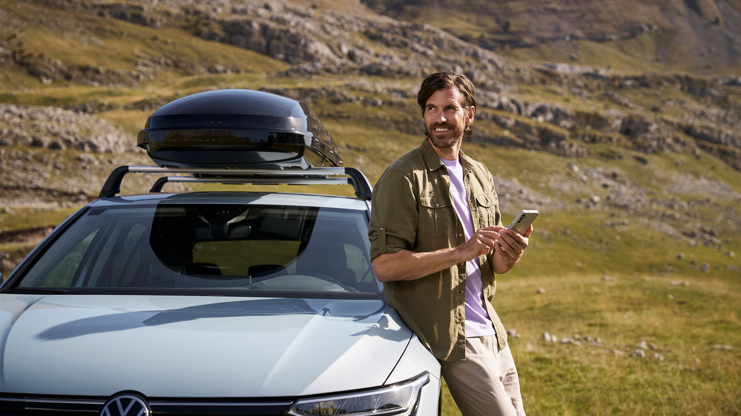 Ein Mann lehnt an einem silbernen VW Modell an und schaut in die Ferne, während er sein Handy in der Hand hält.
