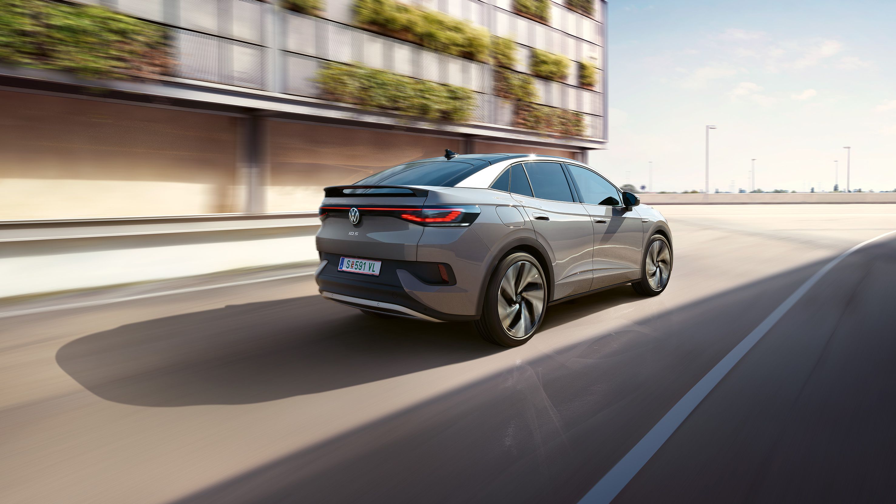 Fahraufnahme eines grauen VW ID.5 von schräg hinten, links ein helles, teilweise grün bewachsenes Gebäude
