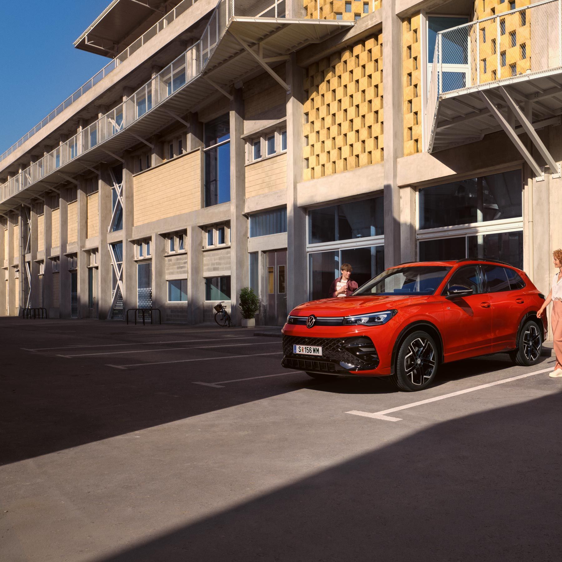 Червен VW Tiguan в профил е паркиран пред сграда, млада жена отива към шофьорското място.