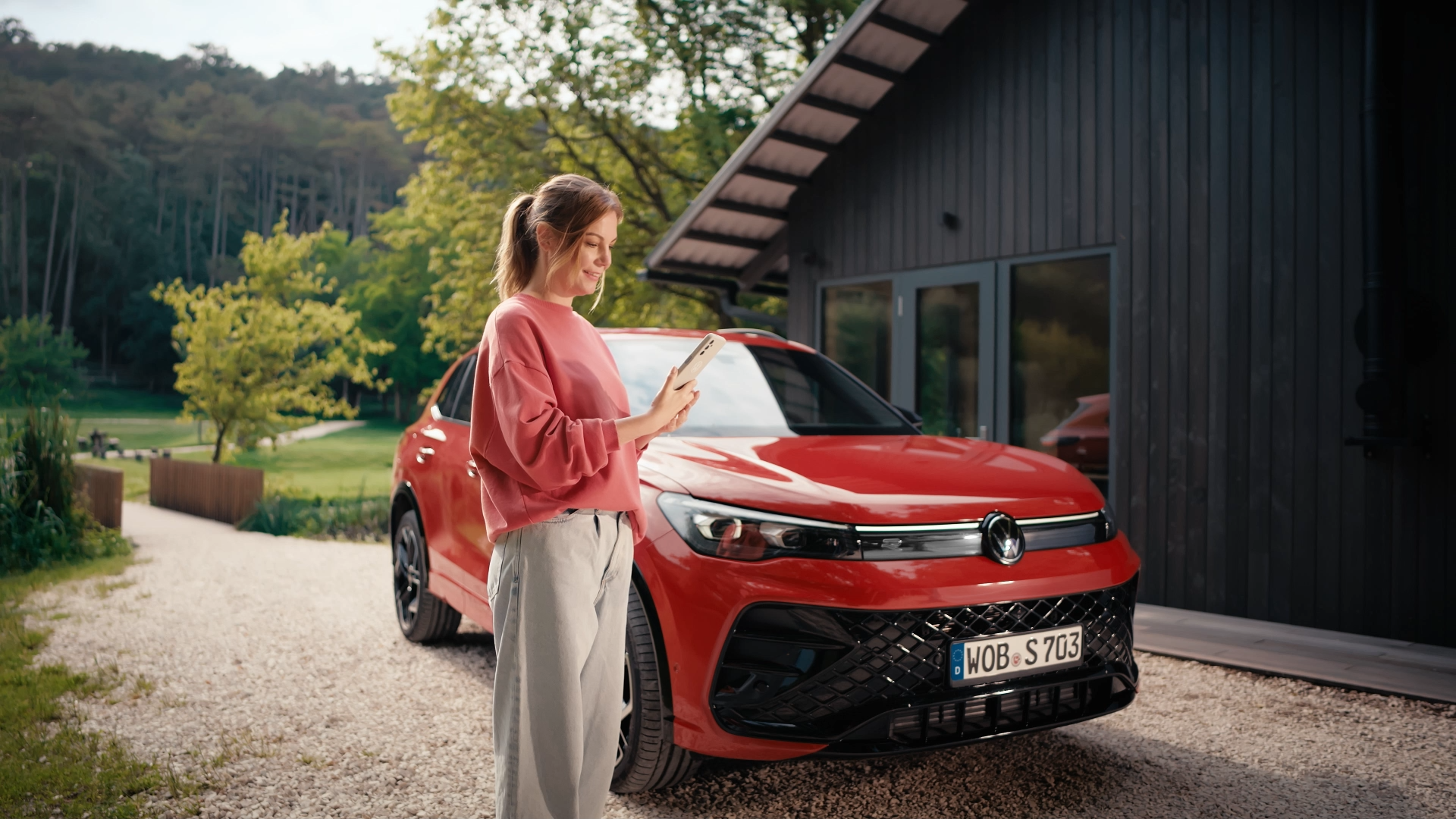 Жена стои пред червения Volkswagen T-Roc и използва своя смартфон пред модерна къща сред природа.
