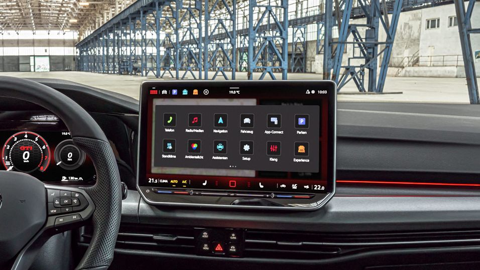 Das Bild zeigt das Cockpit eines modernen Golf GTI. Auffällig ist das große Touchscreen-Bedienfeld und die digitale Instrumententafel. Im Hintergrund ist eine große Industriehalle zu sehen.