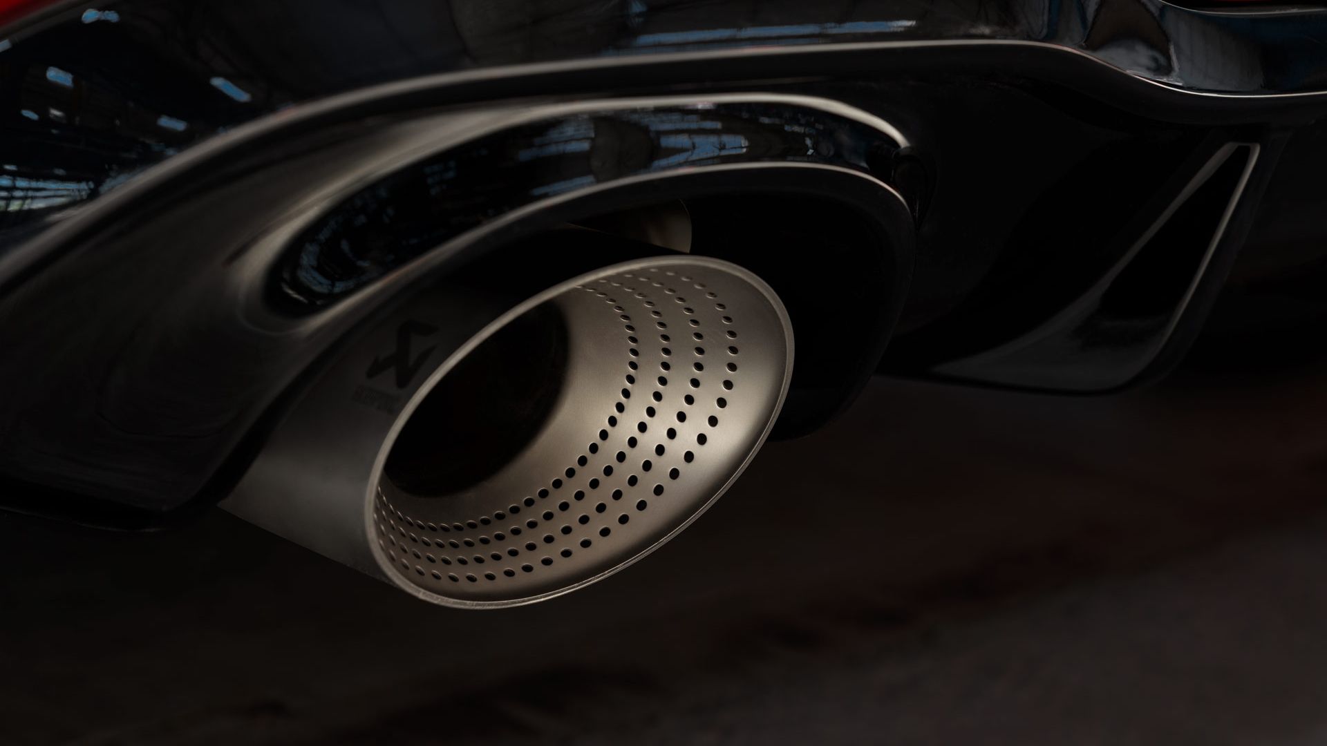 Nahaufnahme eines Autosportauspuffs mit silbernem, perforiertem Endrohr. Das Auspuffrohr gehört zu einem roten Golf GTI, und die glänzend schwarze Karosserie ist leicht zu erkennen. Im Hintergrund sind verschwommene Strukturen eines Gebäudes sichtbar.