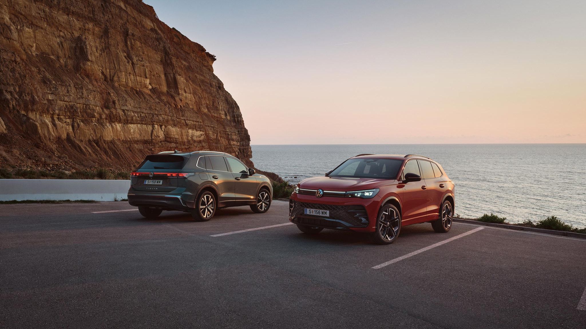 Два нови Volkswagen Tiguan – син и червен – паркирани до скалист бряг с изглед към морето по време на залез.