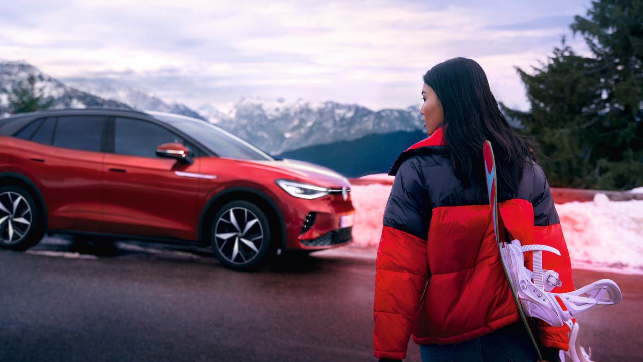 Страничен изглед на червен VW ID.4 GTX на планински фон, на преден план жена гледа към автомобила