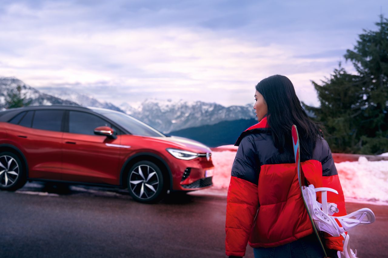 Страничен изглед на червен VW ID.4 GTX на планински фон, на преден план жена гледа към автомобила